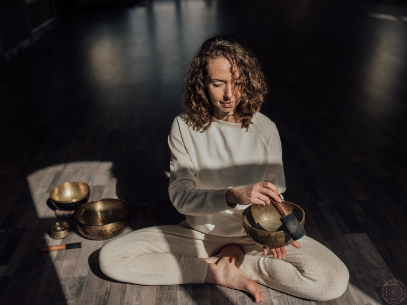 Woman playing a singing bowl with her eyes closed in dim light, symbolizing inner healing and introspection during the Dark Night of the Soul. 