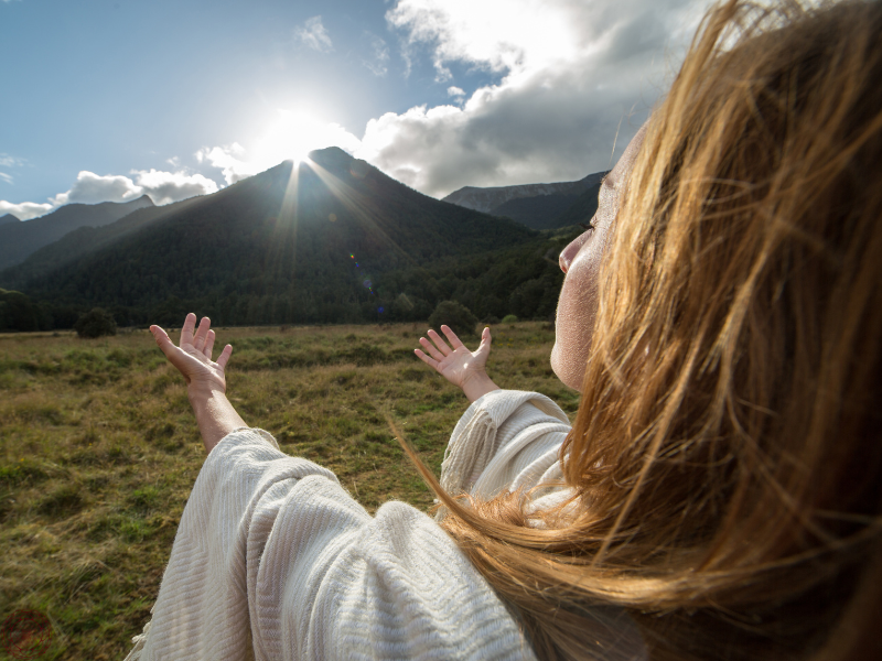 Woman with eyes closed, extending her hands outward towards a mountain, symbolizing spiritual connection and awakening after the Dark Night of the Soul.