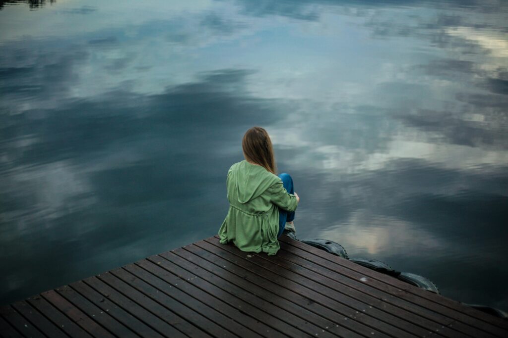 Woman sitting on the edge of a dock in a green jacket, symbolizing solitude and contemplation during the Dark Night of the Soul.