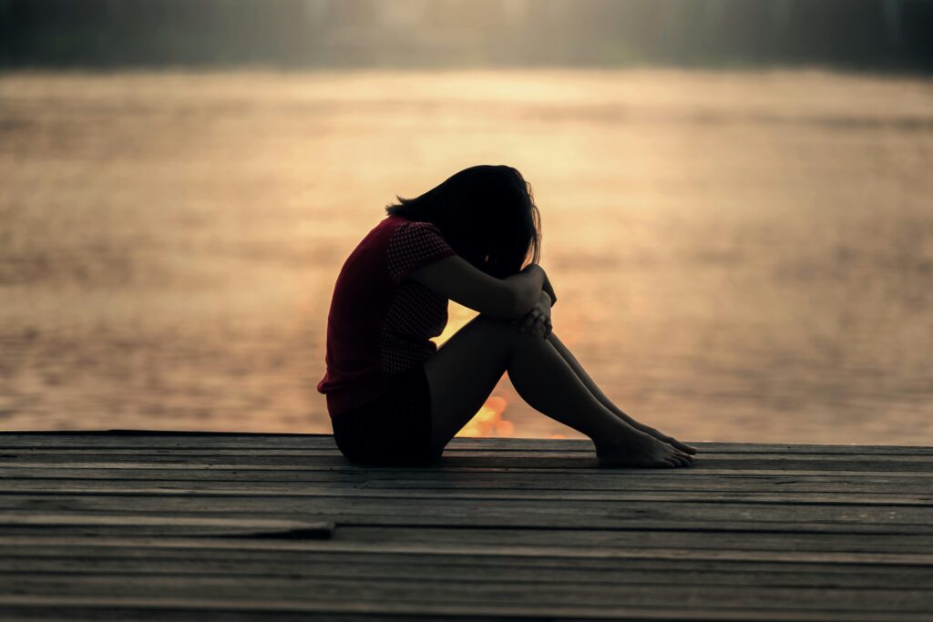 Woman sitting on a dock with her head resting on her knees in dim light, embodying deep emotional reflection during the Dark Night of the Soul.