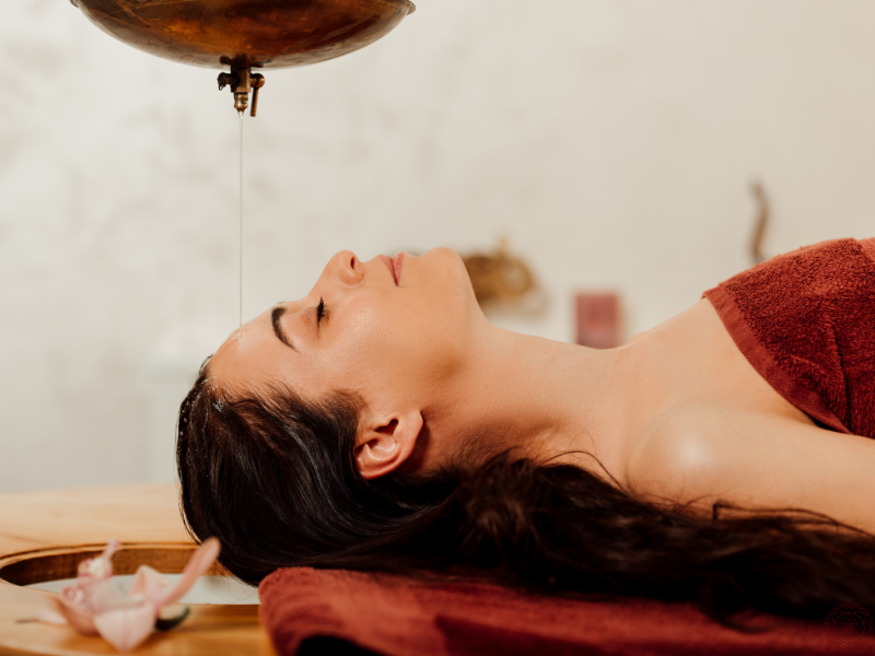 A woman lying down with a towel wrapped around her body, receiving a Shirodhara treatment with oil steadily pouring onto her forehead.