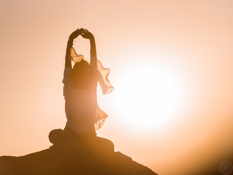 A woman sitting on a mountain top with her hands raised above her head in a meditative gesture as the sun sets behind her.