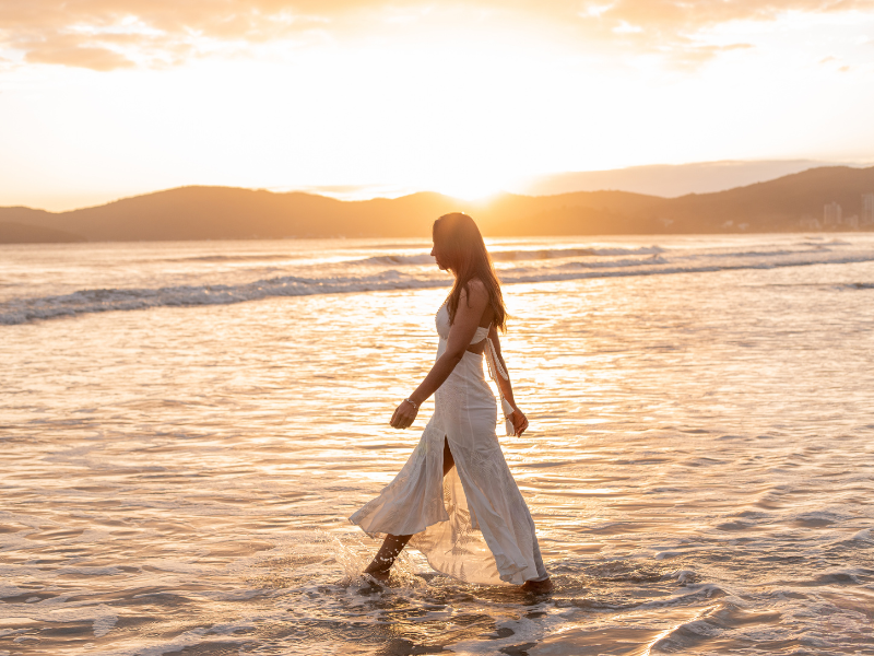 A woman in a flowing white dress walking slowly along the ocean shore at sunset, reflecting the journey of self-realization and divine harmony.