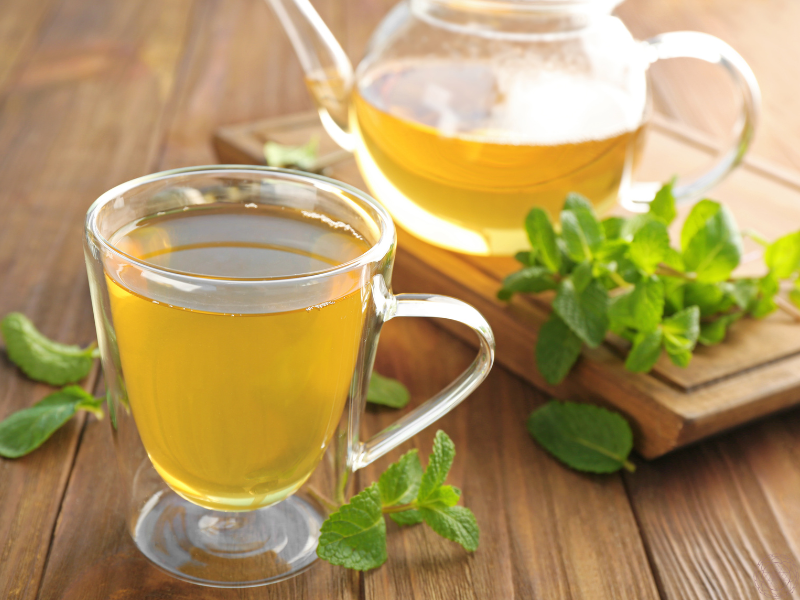 A steaming glass mug and kettle of lemon balm tea on a wooden board with fresh leaves beside them, representing its calming and digestive benefits.