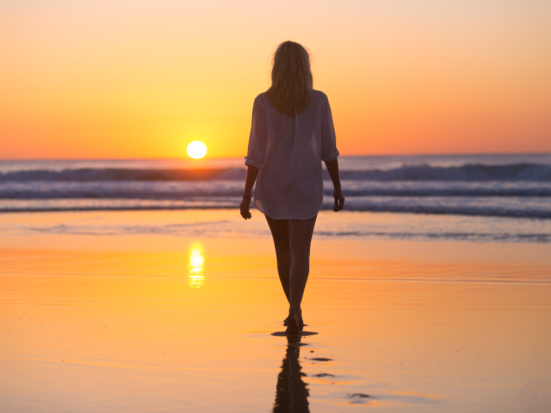 A woman in a white dress walking toward the ocean shore at sunset, symbolizing the introspective and soulful nature of love.