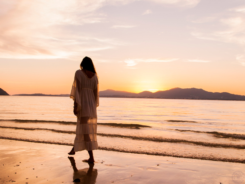 A woman in a flowing white dress walking along the shoreline at sunset, symbolizing the journey of the soul merging with the divine.