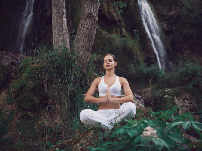 A woman meditating near a waterfall in a lush forest with hands in anjali mudra, embodying tantra’s deep connection to nature, reverence, and inner stillness.