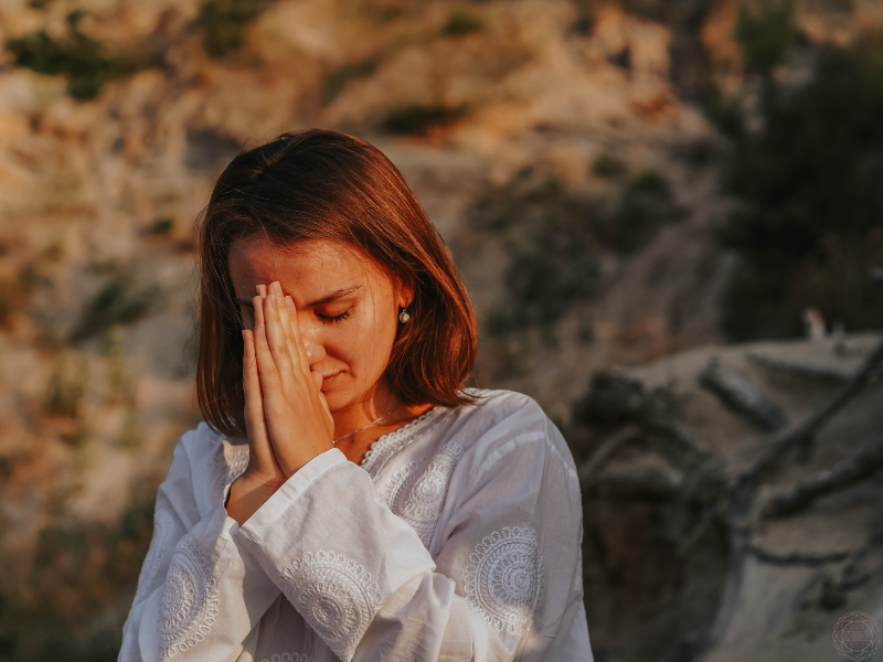 A peaceful woman in a white dress with her eyes closed and hands in prayer at her forehead, reflecting the mindful, heart-centered approach of Ayurveda.