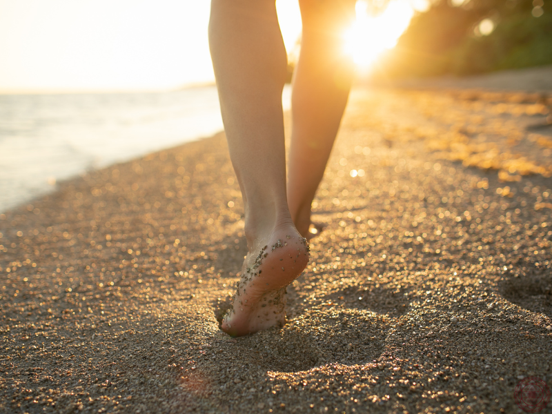 A close-up of bare feet walking along the beach as the sun sets, representing the calming and restorative health benefits of walking.