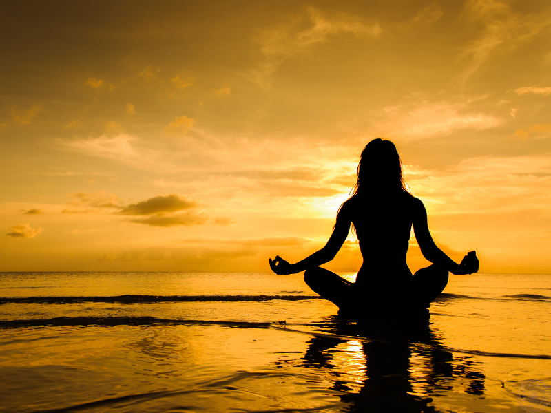 The silhouette of a woman meditating on a beach at sunset, embodying the quiet integration of light frequency into the physical body.