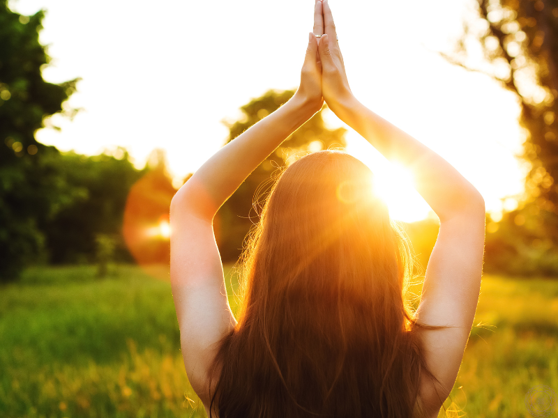 The back of a woman with her hands in prayer position above her head, sunlight streaming through her arms.