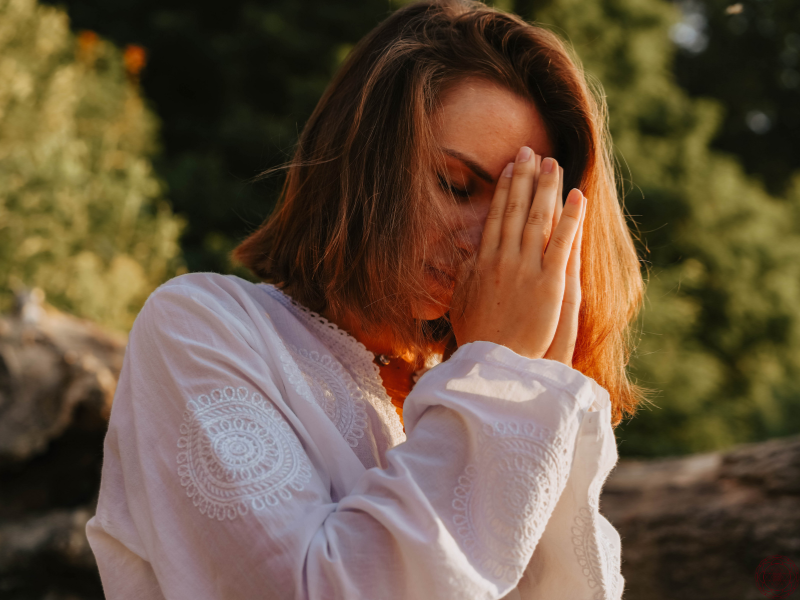 A woman in a white dress with her hands in anjali mudra at her forehead, representing reverence, spiritual devotion, and the sacredness of Kundalini awakening.