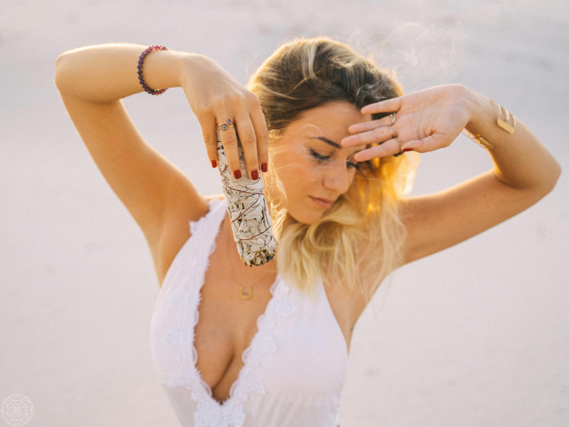 A woman in a white dress dancing with her eyes closed while holding a bundle of sage, symbolizing the awakening and movement of Kundalini energy.