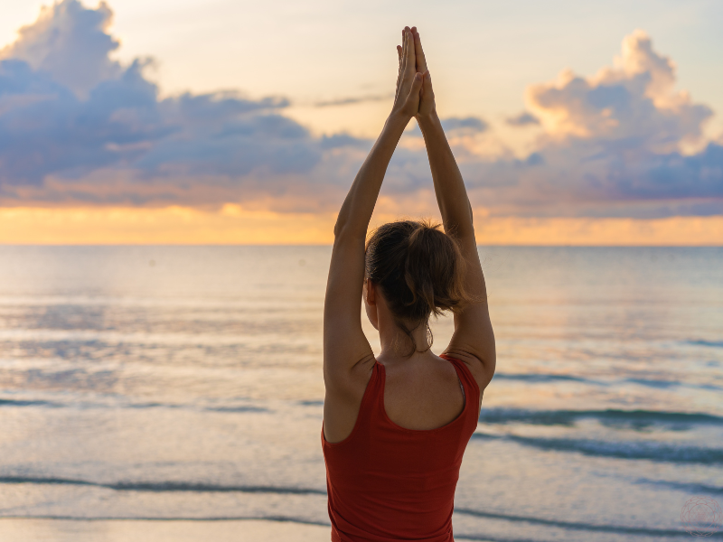 A woman in a red tank top standing at the ocean shore with hands raised above her head, symbolizing the full expression and liberation that comes with a Kundalini awakening.