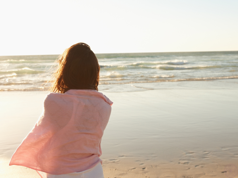 The backside of a woman standing on the ocean shore, gazing at the water, symbolizing introspection, emotional overwhelm, and the quiet weight of living in fear.