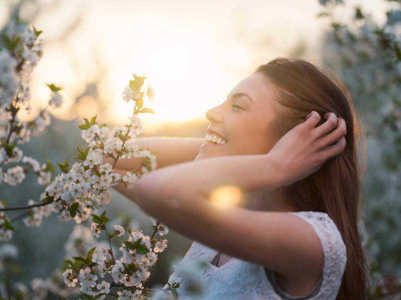 A woman smiling with her eyes closed beside blooming white flowers at sunset, evoking the peace and joy that come from living a purpose-driven life.