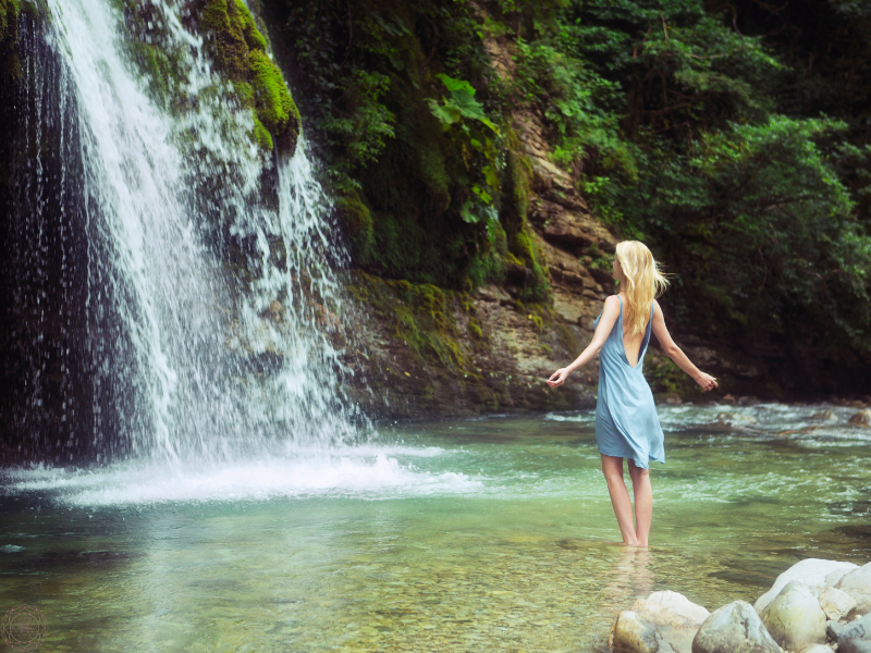 A blonde woman in a blue dress standing in a pool of water with arms outstretched, gazing at a waterfall-symbolizing surrender, alignment, and awe during synchronous moments.