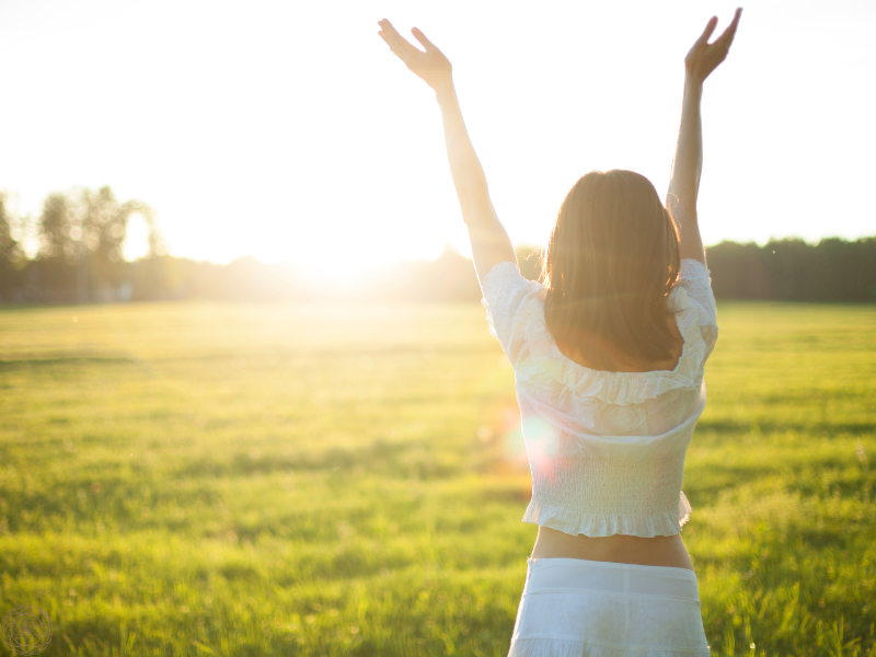 A woman in a white outfit standing in a grassy field with her arms raised above her head, symbolizing connection to breath, openness, and grounded energy in pranayama practice.