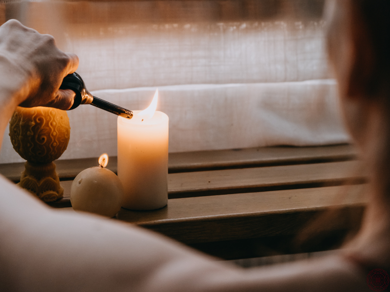 A close-up of a woman gently lighting a candle resting on a wooden bench, symbolizing the quiet ignition of inner awareness through the senses.