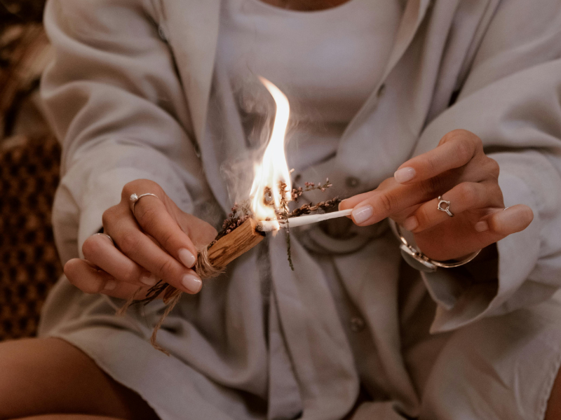 A close-up of a woman burning a bundle of sage, the smoke curling gently upward, symbolizing purification and the lifting of illusion.