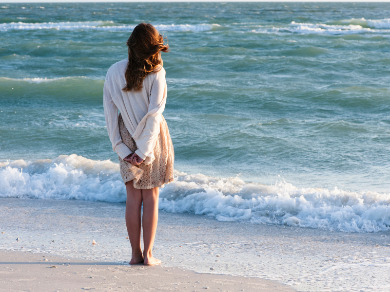 The back of a woman wearing a dress as she stands on the beach, gazing out at the ocean, symbolizing openness and trust.