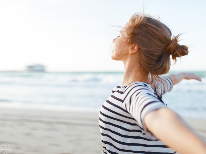 Woman standing on the beach with eyes closed and arms outstretched, symbolizing peace in the face of uncertainty.