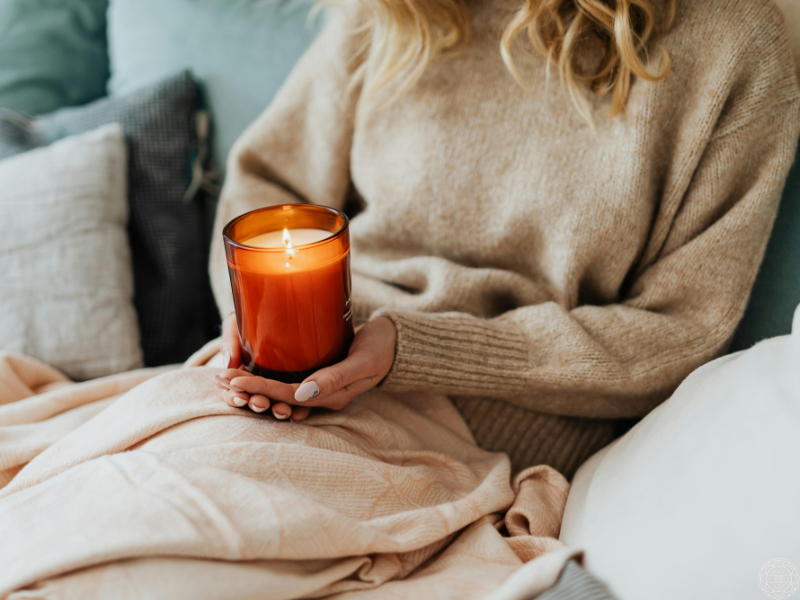 Close-up of a woman sitting in a cozy spot, wearing a sweater, and holding a glowing candle, symbolizing awakened doing.
