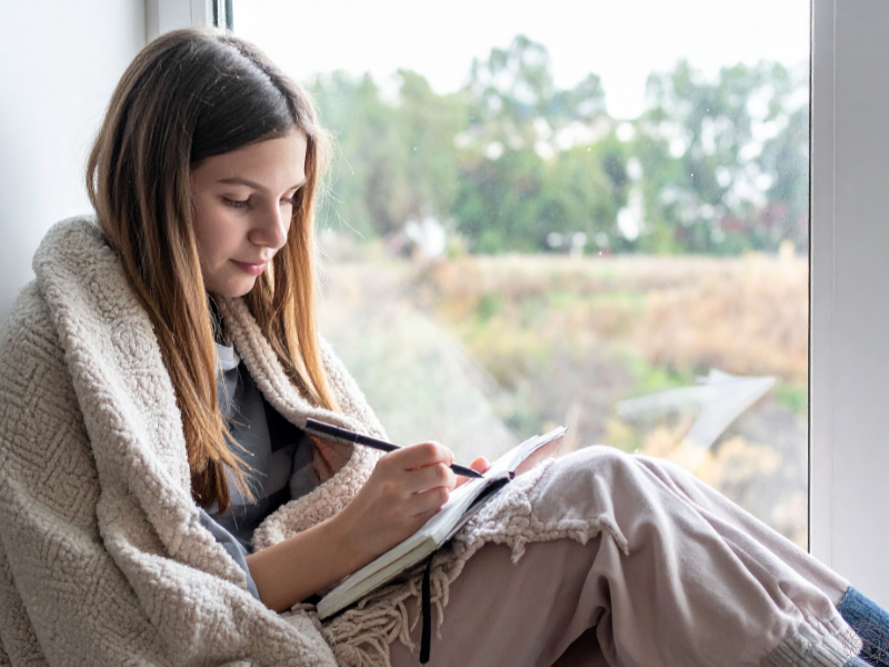 Woman wrapped in a blanket journaling in a cozy spot, reflecting inner awareness and awakened doing.