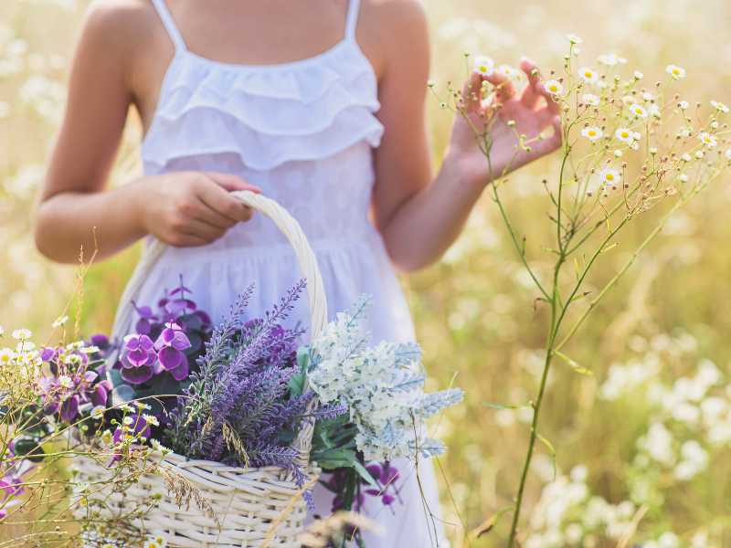 Woman in a white dress holding a basket of flowers while standing in a flower field, embodying presence and awakened doing.