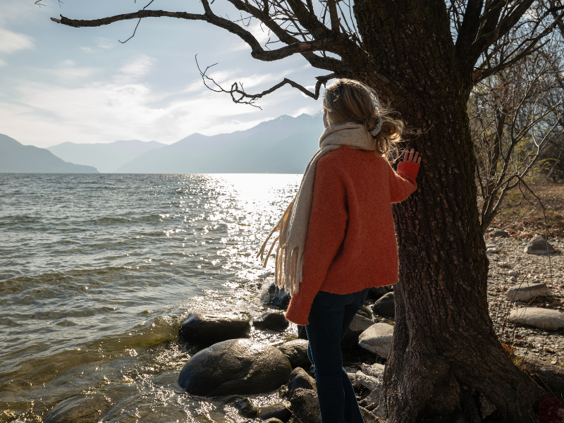 Woman leaning against a tree and gazing at a lake in contemplation, reflecting awareness of the pain-body.