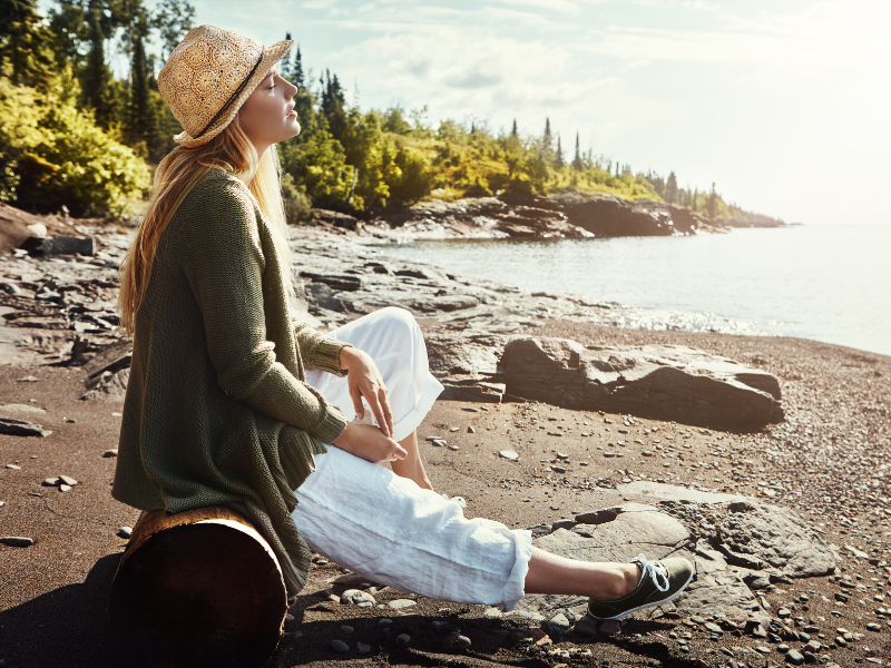 Woman sitting on a log at the beach with her eyes closed, symbolizing stillness and release from the pain-body.