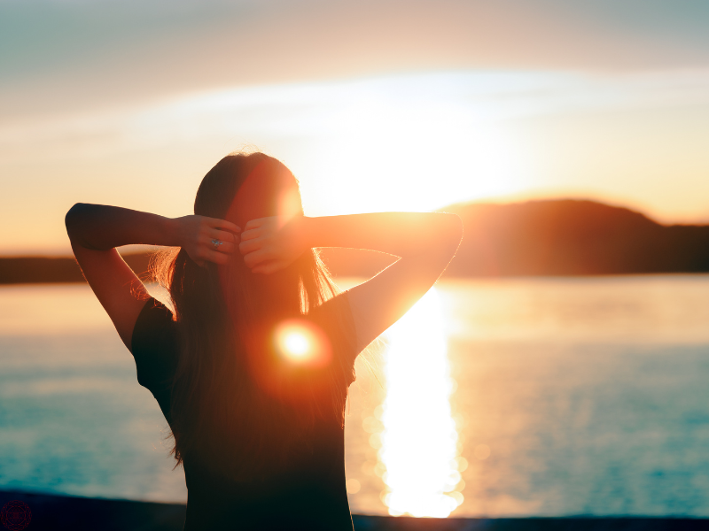 Woman gazing at a large body of water with her hands behind her head as the sun sets, reflecting peace and higher perspective in 5D awareness.