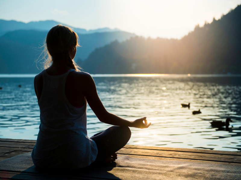 Woman meditating peacefully on a dock overlooking a lake at sunset, symbolizing deep self-reflection and emotional balance through shadow work.