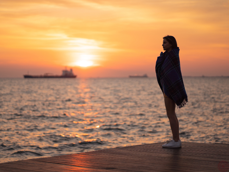 Woman standing on a dock wrapped in a blanket at sunset, symbolizing comfort and acceptance after emotional release through shadow work.