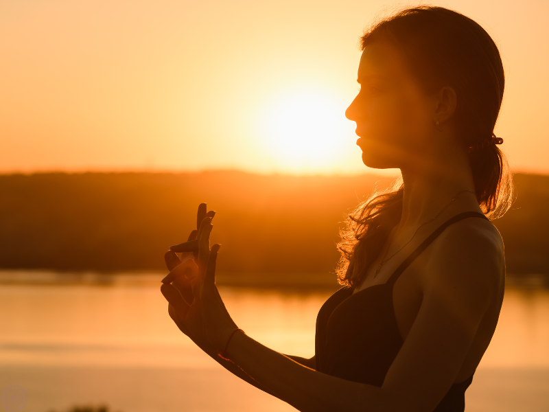 Woman holding up a hasta mudra as the sun sets around her, symbolizing spiritual connection and energy flow.