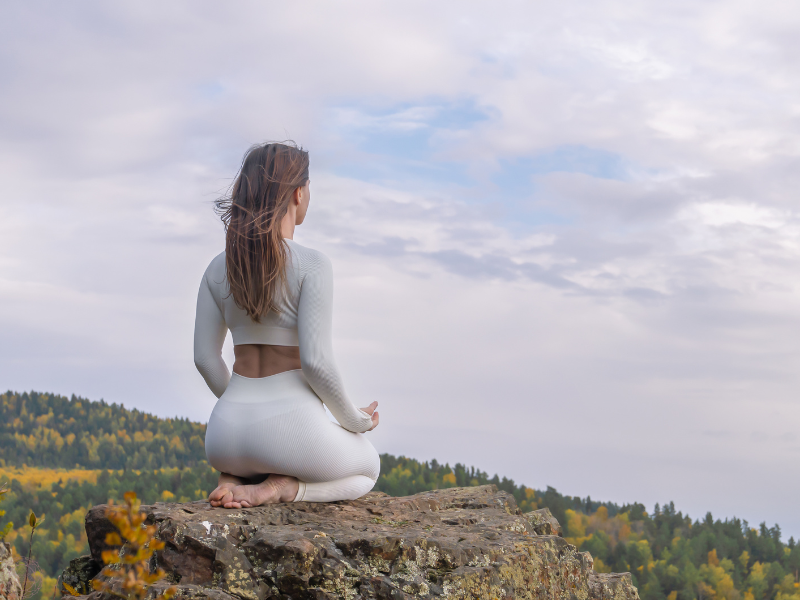 Back of a woman sitting on a rock in a meditative position, symbolizing deep contemplation and higher states of consciousness.