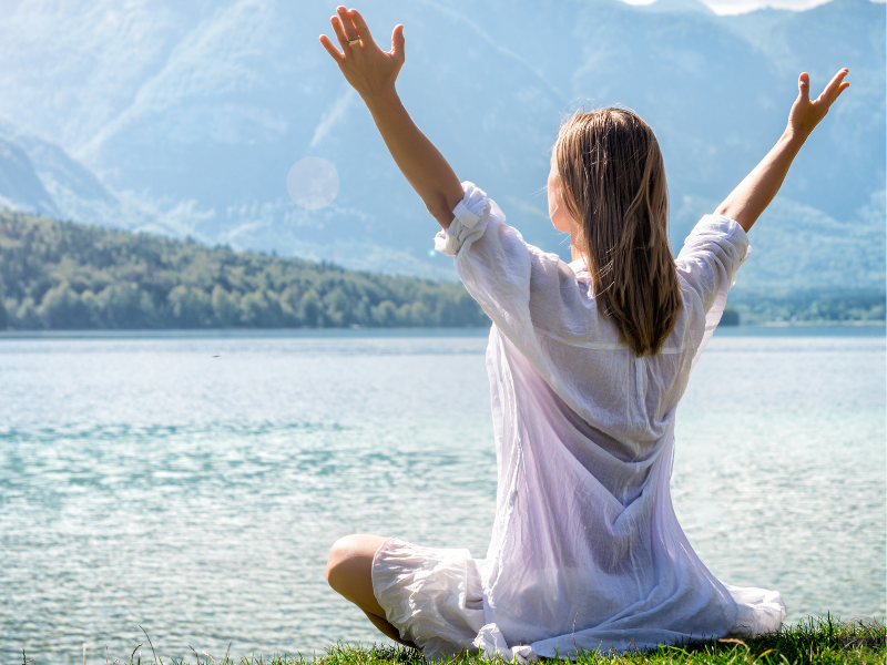 Woman in a white dress sitting near a lake with her hands raised above her head, connecting with Divine consciousness and inner stillness.