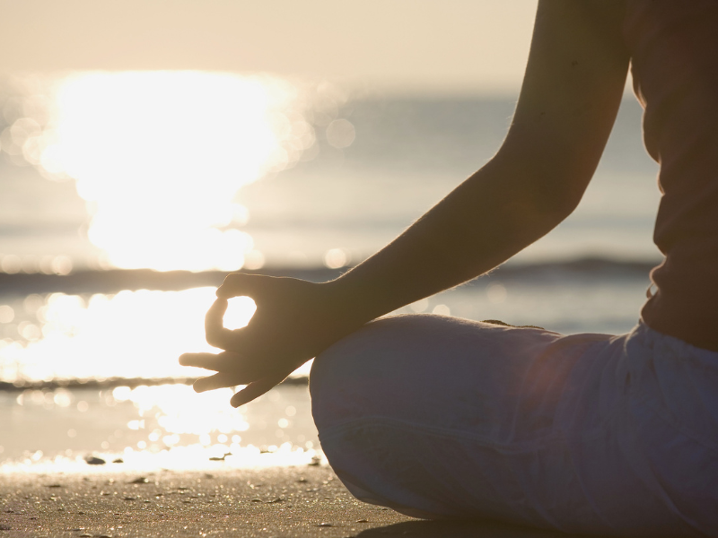 Close-up of a woman’s hands in Gyan Mudra as she meditates on the beach during sunset, symbolizing clarity and alignment with a higher timeline.