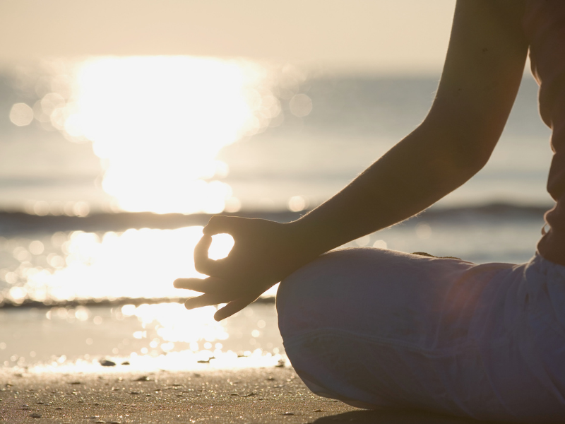 Side view of a woman meditating on a beach with her hand in a mudra, representing the activation and harmonization of nadis through meditation and yogic practices.
