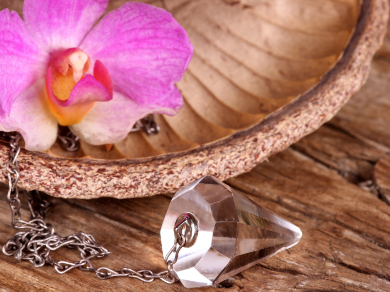Close-up of a crystal pendulum with a pink flower in the background, symbolizing duality and the balance between opposing forces.