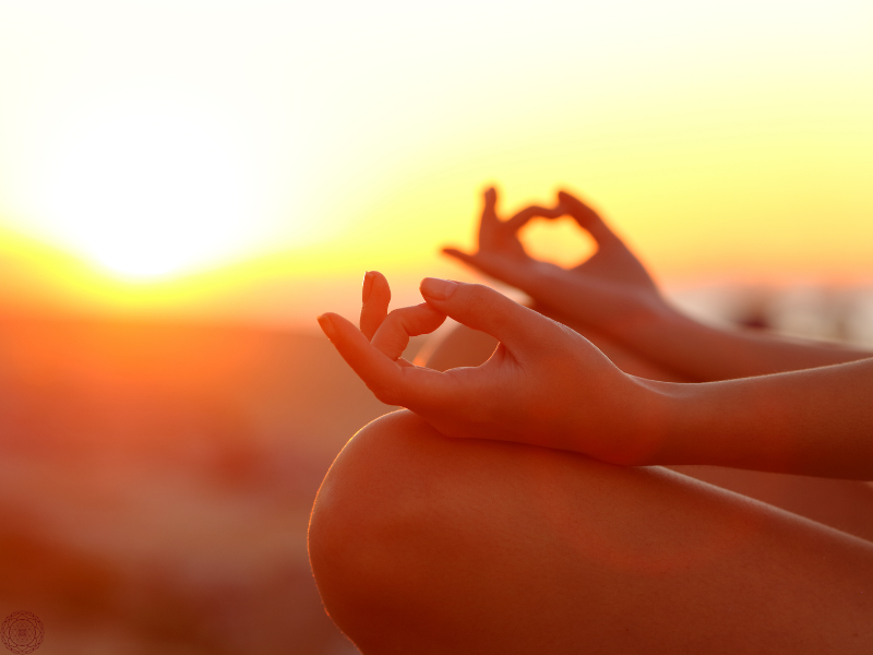 Close-up of a woman’s hands in a mudra with the sun setting behind her, reflecting the contemplative practices described in the Yoga Sutras.